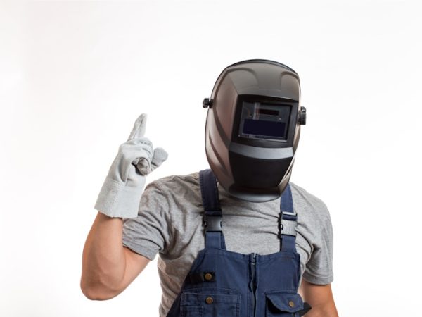 Welder sedang menyiapkan peralatan kerja welder lengkap dengan helm, sarung tangan, dan mesin las di workshop.