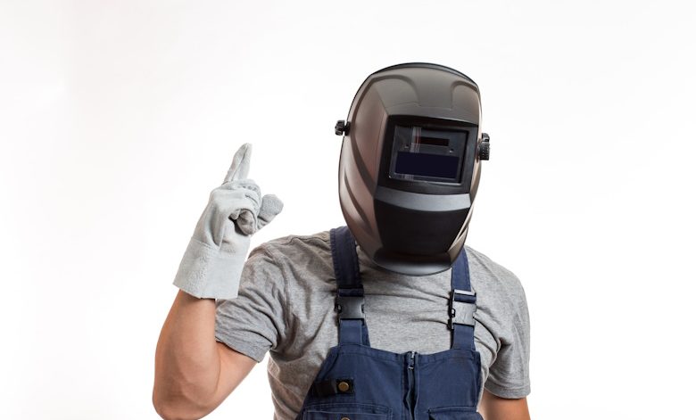 Welder sedang menyiapkan peralatan kerja welder lengkap dengan helm, sarung tangan, dan mesin las di workshop.